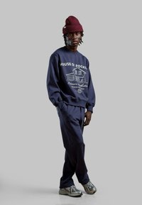 Felpa navy con stampa grafica bianca e pantaloni jogger abbinati. Il modello indossa un berretto bordeaux e sneakers argentate con una finitura testurizzata.