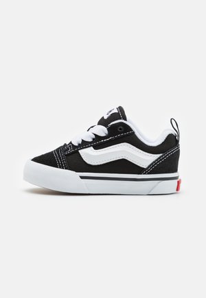 Czarno-białe niskie sneakersy z białymi sznurowadłami, falistym paskiem po boku i białą gumową podeszwą, pokazane z boku na jednolitym tle.