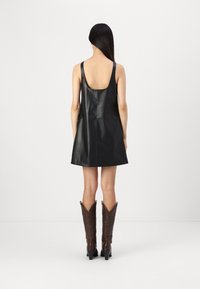 Kvinna med långt svart hår iförd en ärmlös svart läder miniklänning och bruna högklackade cowboystövlar, ståendes mot en enkel vit bakgrund.