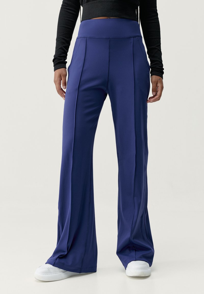 Mörkblå utsvängda byxor i stretchigt material, med hög midja och vertikala sömmar, stylade med vita sneakers.