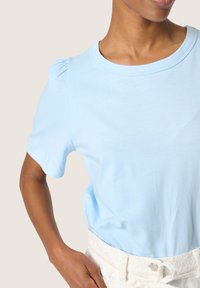 Lyseblå kortærmet t-shirt med rund halsudskæring og samlet skulderdetalje, lavet af blødt stof. Båret med hvide teksturerede shorts.
