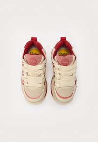 Zapatillas beige y rojas con forma redondeada, tejido texturizado, cuello acolchado y cordones prominentes. Suela amarilla visible con la marca.