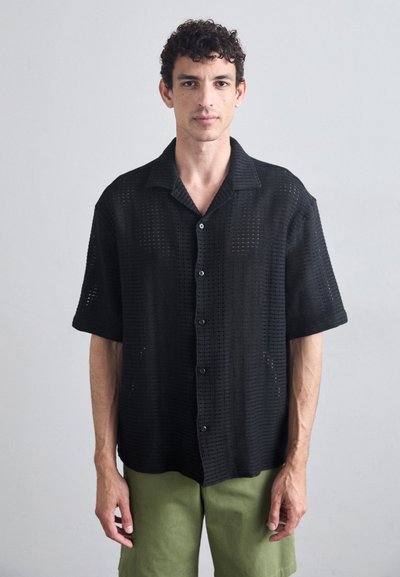 Chemise noire à manches courtes, à boutons, avec un design en maille ouverte texturée. Présente un col classique et une coupe décontractée, associée à un pantalon vert clair.
