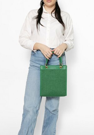 Donna che indossa una camicia bianca e jeans azzurri, che tiene una borsa verde testurizzata con dettagli ad anello dorati sulle maniglie.