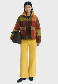 Chaqueta de forro polar con un diseño de patchwork en naranja, verde y marrón, que cuenta con dos bolsillos frontales y un cuello alto. Combinada con pantalones amarillos de canalé.