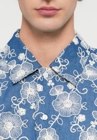 Κοντινό πλάνο του κάτω μέρους του προσώπου και του λαιμού ενός ατόμου που φοράει μπλε πουκάμισο με λευκή κεντημένη λουλουδάτη διακόσμηση και γιακά με κουμπιά.