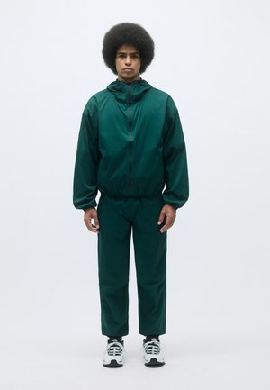 Zaļa, viegla treniņtērpu komplekts no gludas auduma, kas ietver rāvējslēdzēja jaku ar kapuci un elastīgām piedurknēm, kopā ar atbilstošām saplacinātām biksēm.