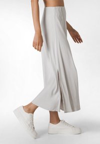 Jupe longue et fluide gris clair avec une fente sur le côté, fabriquée en tissu doux. Associée à des baskets blanches texturées avec une semelle compensée.