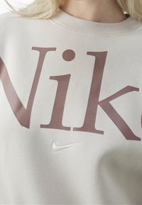 Světlý béžový svetr s velkým bordovým nápisem "Nike" a malým bílým logem Nike swoosh. Hladká textura, žebrovaný výstřih.