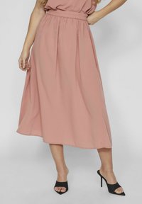 Femme portant une jupe midi rose poussiéreuse avec une taille élastique et des mules ouvertes à talons hauts noires, se tenant devant un fond uni.