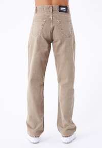 Pantaloni in denim beige con taglio dritto, dotati di tasche posteriori e di un'etichetta nera con logo in vita. Tessuto testurizzato, design casual.