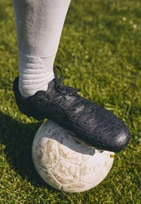 Svarte fotballsko med en teksturert, sprukket design, snørt på en fot over en skitten hvit fotball på gress. Hvit ankelsokk synlig.