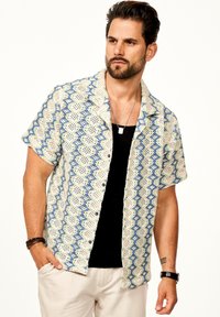 Kurzarmhemd aus Häkelstrick mit blau- und grünem Zickzackmuster auf einem cremefarbenen Hintergrund; verfügt über schwarze Knöpfe und einen kragenartigen Ausschnitt.