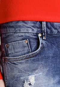 Vaqueros de denim en azul claro con un efecto desgastado, que presentan un bolsillo frontal con remaches plateados y costura visible, junto a una blusa roja a la altura de la cintura.