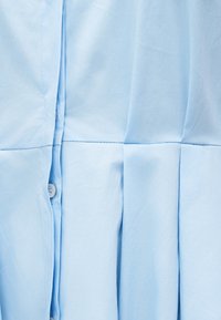 Υφασμάτινο ένδυμα ανοιχτού μπλε χρώματος με οριζόντιο ράψιμο και κάθετες πιέτες, με μερικώς ορατό κουμπί και κουμπότρυπα.