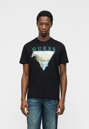 Man draagt een zwart Guess T-shirt met een tropisch strandtafereel binnen een driehoek en een blauwe spijkerbroek, staand tegen een effen lichte achtergrond.