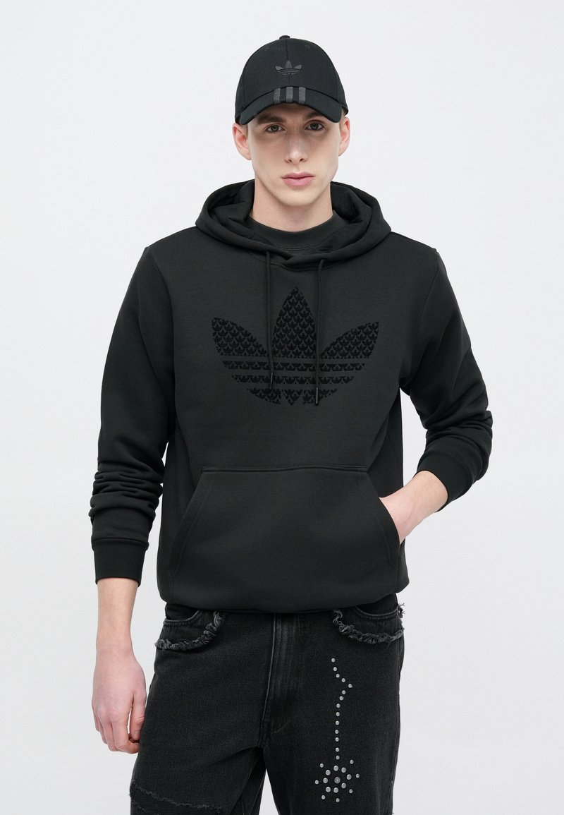 Black hoodie with textured black Adidas logo, front pocket, adjustable drawstrings; black cap with logo and three stripes. Black denim jeans with studs.