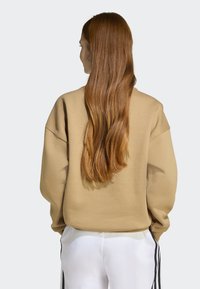 Beige oversized sweatshirt med sänkt axelparti, visad bakifrån, matchad med vita byxor med svarta sidostråk. Långt hår synligt.