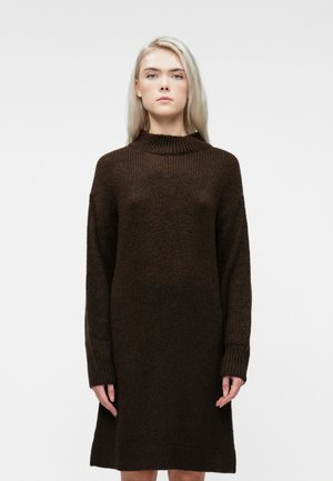 Young woman with straight blonde hair wears a long-sleeve, dark brown knitted sweater dress, standing against a white background.