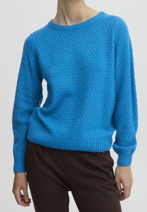 Blå, tekstureret striktrøje med rund halsudskæring, båret med mørkebrune bukser, vist på en stående person fra nakke til hofter.