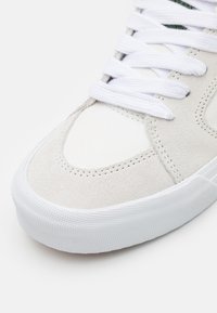 Chaussure de sport blanche avec une tige en daim et toile, bout rond, semelle en caoutchouc texturé et lacets blancs plats sur une languette rembourrée.