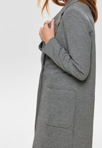 Donna che indossa un lungo cappotto grigio con una tasca frontale, tenendo un lato del cappotto vicino al colletto, su uno sfondo chiaro e uniforme.