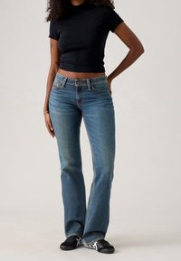 Femme portant un t-shirt court noir, un jean bootcut bleu et des baskets noires, debout avec une main sur la hanche devant un fond uni.