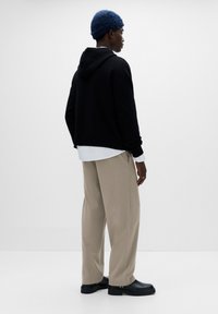 Fekete kapucnis pulóver, fehér ing alatta, bézs, széles szárú nadrág és fekete cipő. Kék, texturált kötött sapka. Minimalista háttér.