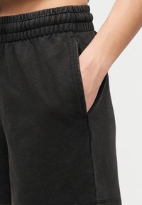 Άτομο με το χέρι στην τσέπη σορτς με ελαστική μέση σε σκούρο γκρι χρώμα, μπροστά σε απλό ανοικτό φόντο.