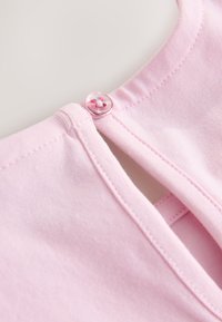 Detail světle růžového výstřihu oděvu s malým kulatým růžovým knoflíkem a zapínáním na látkovou smyčku.