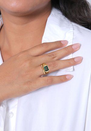 Goldring mit einem rechteckigen grünen Stein, glatte Oberfläche, minimalistisches Design, getragen an einer Hand mit hellrosa Nägeln vor einem weißen Hemd.