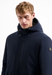 Donkerblauwe hoodie gemaakt van gladde stof, met een voorzak met rits en een hexagonaal logo op de mouw.