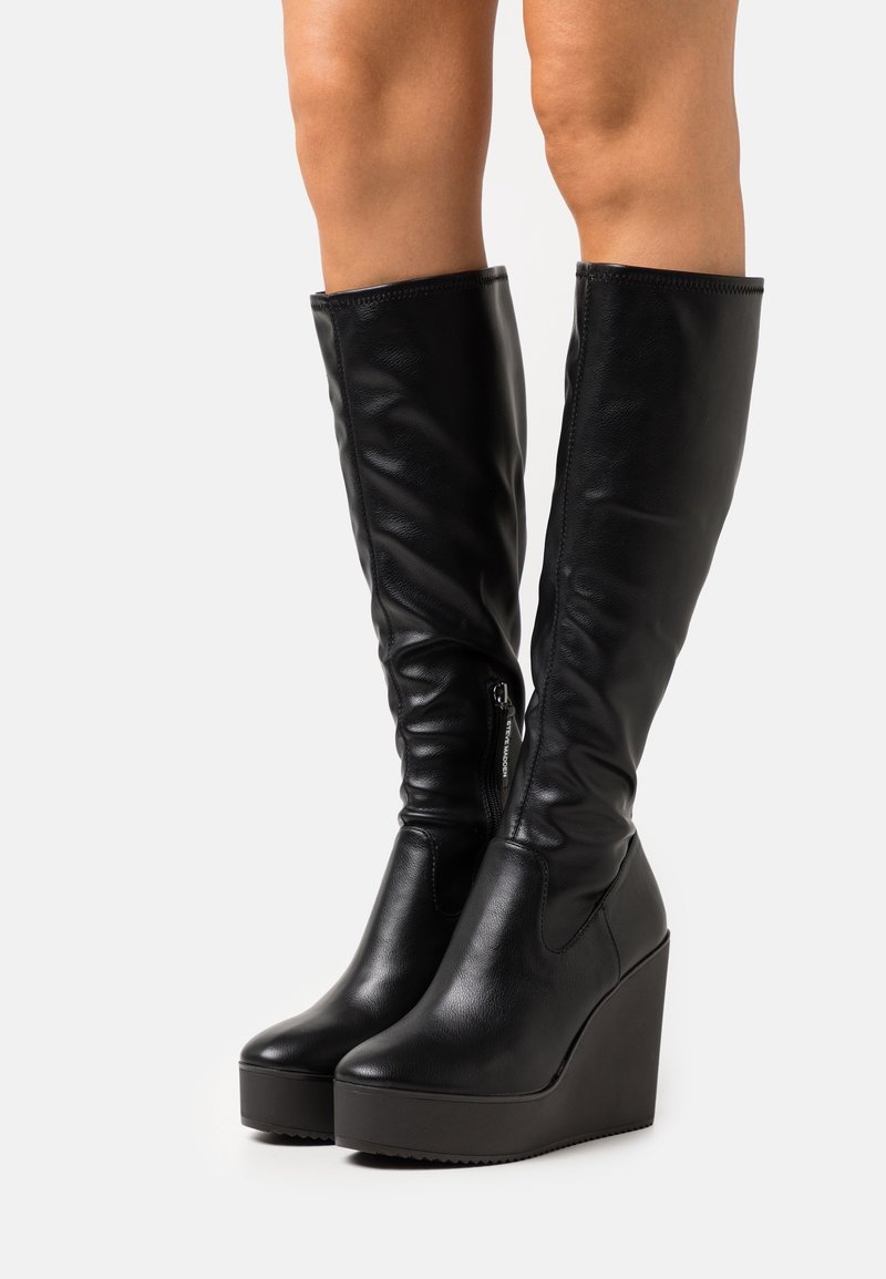 Black faux leather knee-high boots with a slouchy design, pointed toe, and a chunky wedge sole. Features a side zipper for easy wear.
