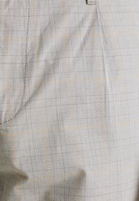 Harmaa kangas, jossa on ruudullinen kuvio ohuilla oransseilla viivoilla. Sileä pinta, räätälöity istuvuus ja hienovarainen kiilto. Näkyviä saumojen yksityiskohtia.