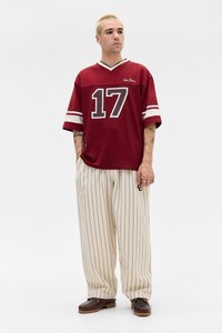 Maglia rossa con strisce bianche e grafica "17"; abbinata a pantaloni beige a righe larghe e scarpe marroni senza lacci.