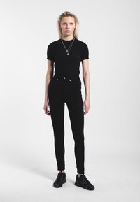 T-shirt aderente nera, jeans skinny neri a vita alta e sneakers neri. Collane d'argento a strati aggiungono un tocco discreto all'outfit.