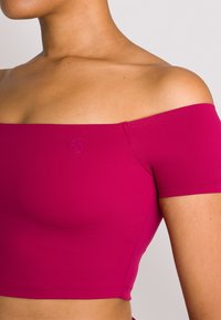 Dopasowany, hiszpański crop top w intensywnym magencie, wykonany z gładkiego, elastycznego materiału. Posiada krótkie rękawy i subtelny detal z logo.