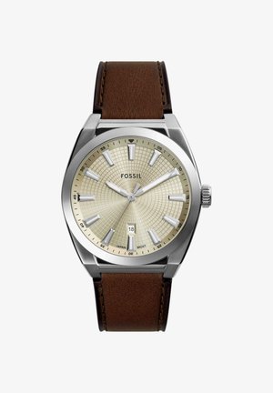 Analog Fossil wristwatch with beige textured dial, silver markers, date display at 6 o'clock, and brown leather strap.