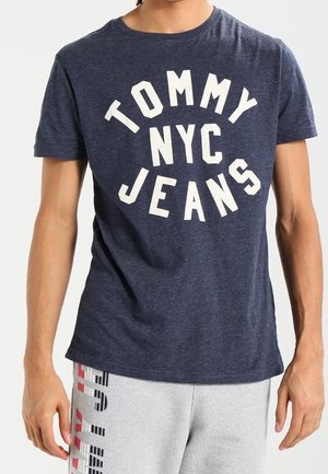 Navy blue cotton t-shirt featuring white text reading “TOMMY NYC JEANS,” with a crew neck and short sleeves. Worn with light grey joggers.