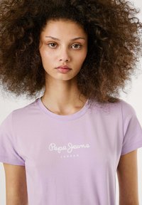 Gaiši violeta T-krekls, kas izgatavots no mīkstas kokvilnas, ar apaļu kakla izgriezumu un īsām piedurknēm, uz kura baltā krāsā ir druka "Pepe Jeans London".