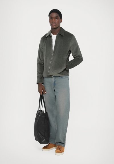 Grå kordjakke med lynlås foran, flad krave. Lyst blå lige ben jeans. Brune sneakers, holder en sort tote bag.