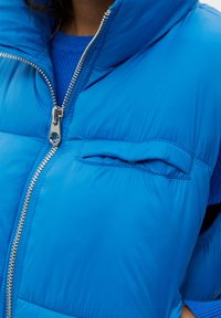 Chaleco acolchado azul con un cuello alto, que presenta una cremallera plateada y un pequeño bolsillo en el pecho. Textura suave y material liviano.