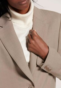 Beige op maat gemaakte blazer over een crèmekleurige coltrui. De blazer heeft drie knoppen op de manchetten en een gladde textuur met subtiele patronen.