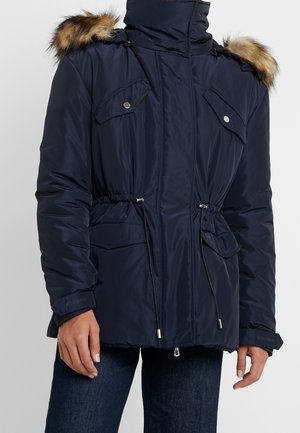 Veste d'hiver bleu marine avec capuche bordée de fourrure, fermeture éclair à l'avant, poches poitrine boutonnées, taille ajustable avec cordon de serrage et poches latérales à rabat.
