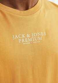 Senf rumena bombažna majica s belim tiskanim logotipom "JACK & JONES PREMIUM" v sredini na prsih, z okroglim ovratnikom.