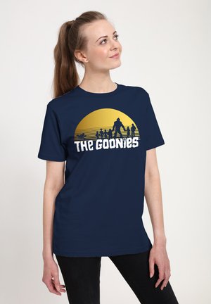 Kobieta z kucykiem, ubrana w granatowy T-shirt z żółtym słońcem i sylwetkami ludzi oraz napisem „The Goonies“.
