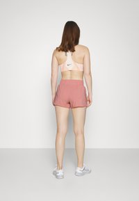Sujetador deportivo en melocotón claro con patrón geométrico, acompañado de pantalones cortos sueltos rosas. La modelo lleva zapatos deportivos blancos.