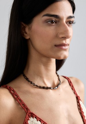 Beaded necklace s kristalima smeđih nijansi, stvarajući kutni dizajn. Nosena na goloj vratu iznad crvenog heklanog topa.