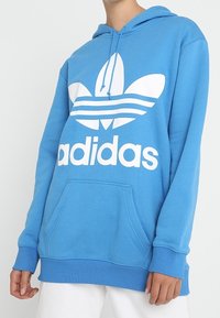 Blå huva sweatshirt i mjukt tyg, med en stor vit Adidas-logotyp och design med tre ränder på framsidan. Framficka ingår.