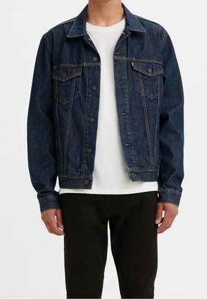 Mann trägt eine dunkelblaue Jeansjacke über einem weißen T-Shirt mit schwarzen Hosen und steht vor einem einfarbigen Hintergrund.